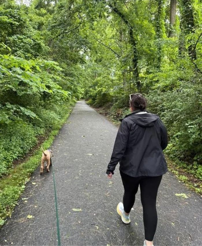Dog walker walking a small dog on a leash along a wooded paved trail in Chantilly, Virginia